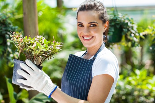 A woman with dark hair pulled back, smiling and wearing a white t-shirt and blue striped gardening apron, holds a small potted shrub with green and purple leaves. She is standing outdoors in a garden setting, surrounded by lush green foliage, including plants, trees, and bushes, suggesting a well-maintained outdoor space. The background features blurred greenery, indicating a sunny day with natural light illuminating the garden. The scene conveys professional gardening care, typical of services such as hedge trimming and landscape maintenance provided by companies like Hedge Trimming Shepherds Bush, supporting local gardening and landscaping needs in the Shepherds Bush area of London.